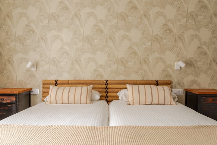 Une chambre confortable équipée de lits jumeaux avec des draps blancs, une tête de lit en bois, des oreillers beiges à rayures et du papier peint d'inspiration tropicale.