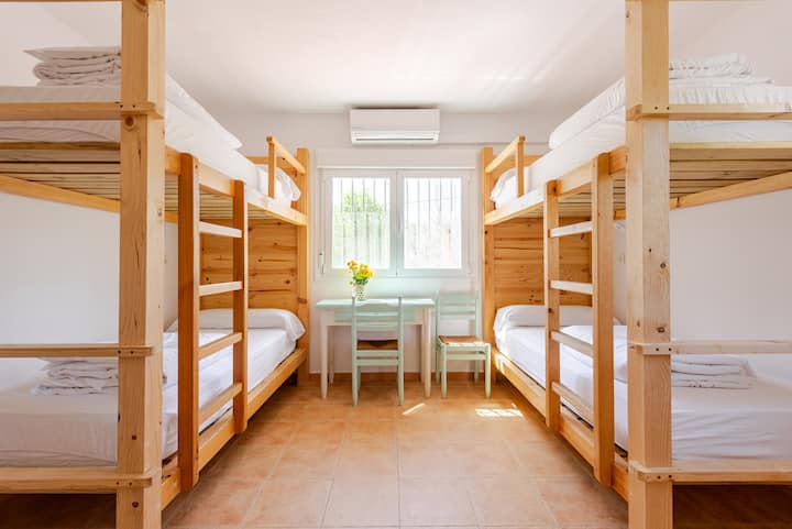 Une chambre minimaliste avec des lits superposés en bois recouverts de draps blancs, un sol carrelé, une petite table et des chaises près d'une fenêtre laissant entrer la lumière naturelle.