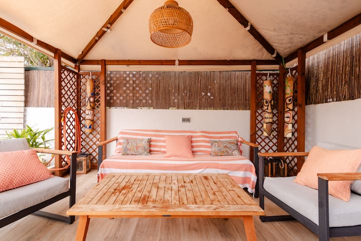 Un salon extérieur chaleureux et accueillant avec un canapé, des fauteuils et une table en bois sous un auvent, décoré de masques tiki et de verdure.