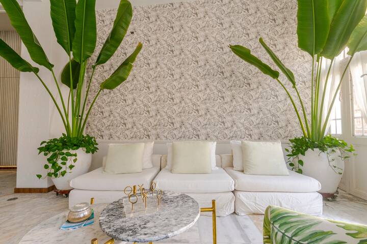 Un salon lumineux et élégant avec un canapé blanc, de grandes plantes tropicales en pot, un papier peint à motifs et une table basse en marbre avec des touches dorées.