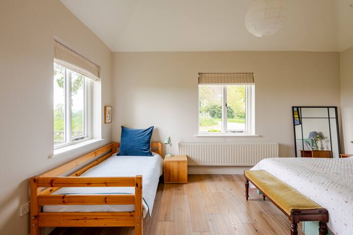 Une chambre lumineuse et aérée avec un lit simple en bois, un lit double et une grande fenêtre donnant sur de la verdure, avec un design minimaliste et confortable.