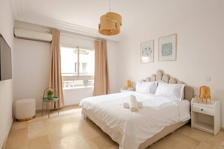 Une chambre décorée avec goût dans des tons neutres, équipée d'un lit queen size, avec des draps blancs et des serviettes empilées, de deux tables de chevet avec des lampes en osier. Une fenêtre laisse entrer la lumière naturelle et des cadres au thème végétal habillent le mur au-dessus du lit.