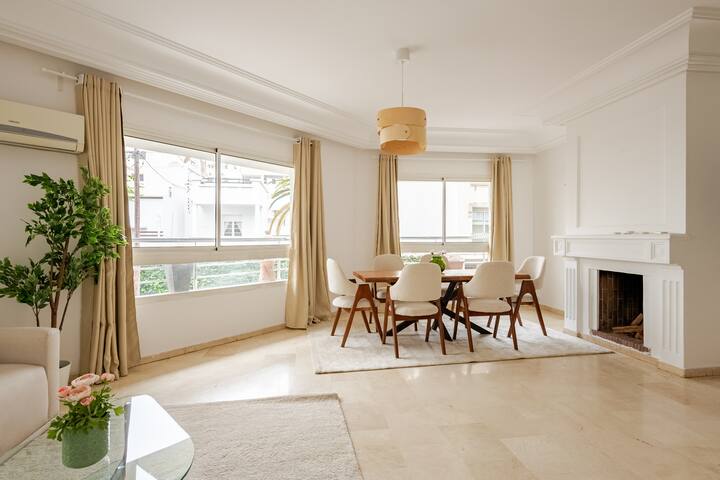 Un espace de vie minimaliste baigné de lumière naturelle, avec une table ronde entourée de chaises blanches, des rideaux beiges et une cheminée blanche.