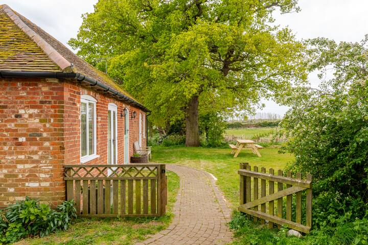 Une scène rurale montrant une maison en briques rouges avec un toit incliné, entourée de verdure et d'un grand chêne. Un chemin incurvé en briques mène à des tables de pique-nique dans le jardin.