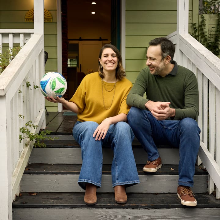 A couple sits, holding a soccer ball on their front porch and smiling.