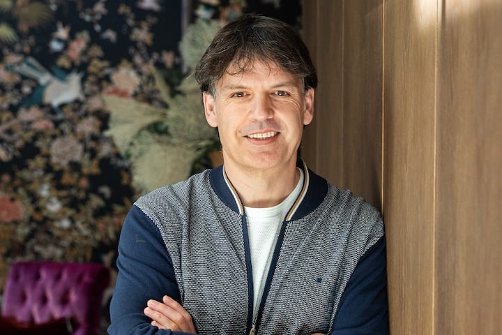 Fernando Morientes, con una chaqueta azul marino y gris, está de pie con los brazos cruzados frente a una pared de madera. Al fondo hay otra pared con un papel pintado de flores. La imagen forma parte de las experiencias de LALIGA que se ofrecen en Airbnb.