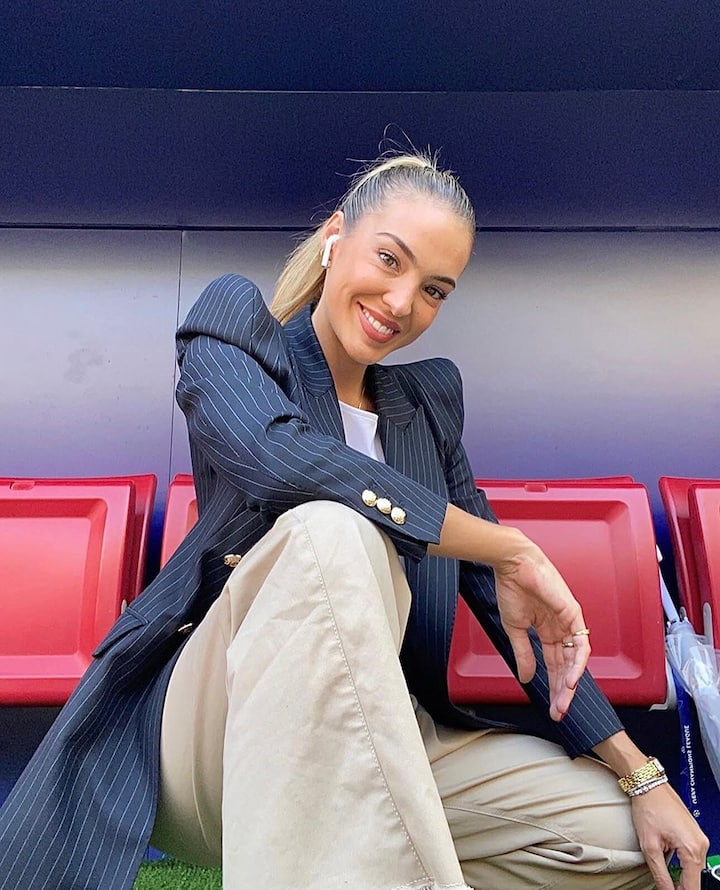 Primer plano de Isabel Forner sentada frente a los asientos de color rojo intenso de un estadio. Está vestida con un atuendo profesional compuesto por una chaqueta de rayas azul marino y unos pantalones beige.