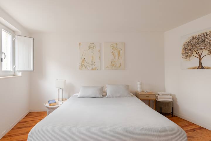 Une chambre confortable et bien éclairée, baignée de lumière naturelle grâce à une fenêtre partiellement ouverte. La chambre a des murs blancs, un plancher en bois, un lit soigneusement fait, des tables de chevet, des croquis de silhouettes humaines au-dessus du lit et un tableau représentant un arbre sur le mur latéral.