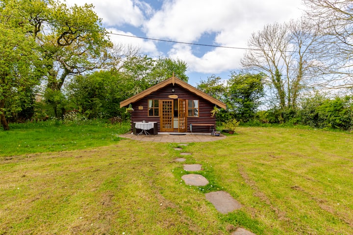 Une cabane en bois rustique située dans un champ paisible et ouvert avec un chemin en pierre menant à l'entrée. La cabane est bordée de grands arbres sous un ciel nuageux.
