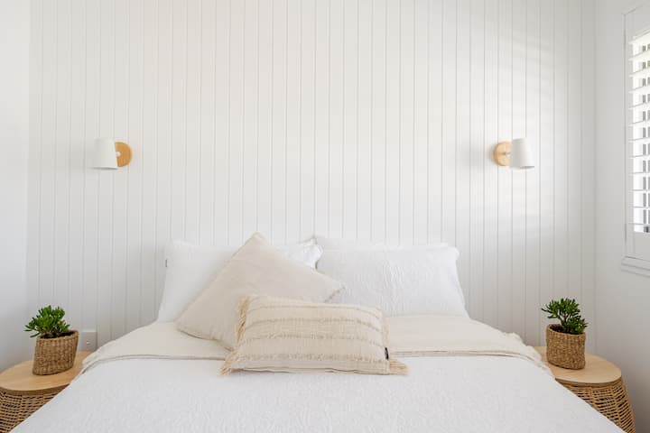 Une chambre minimaliste avec du lambris blanc, un lit confortable décoré de coussins et de touches chaleureuses comme des tables de chevet en osier surmontées de plantes vertes.
