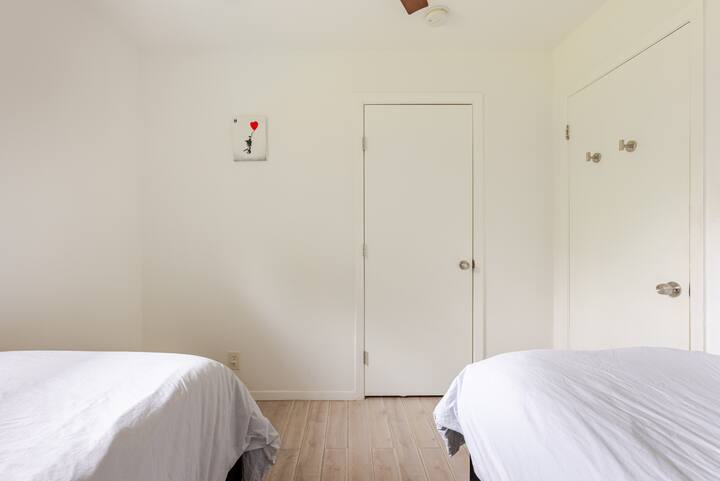 Une chambre propre et aérée avec deux lits simples, des murs blancs, des sols en bois clair et un petit cadre représentant un ballon rouge au-dessus de la porte.