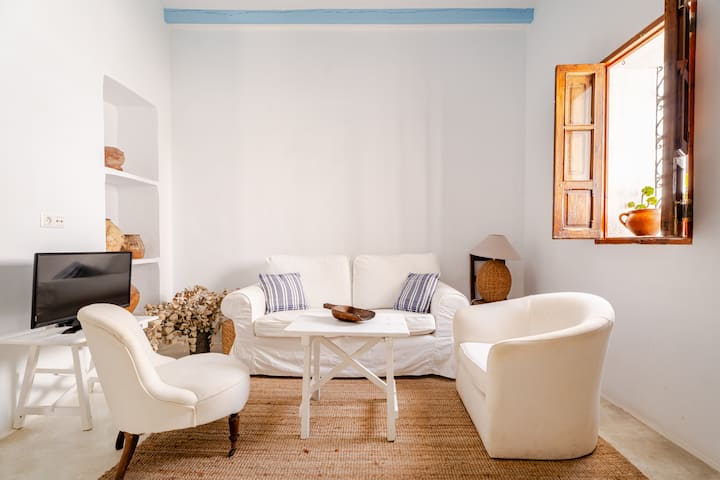 Coin salon confortable avec des meubles blancs, une télévision, un tapis tissé et une fenêtre avec une plante en pot et la lumière naturelle du soleil.