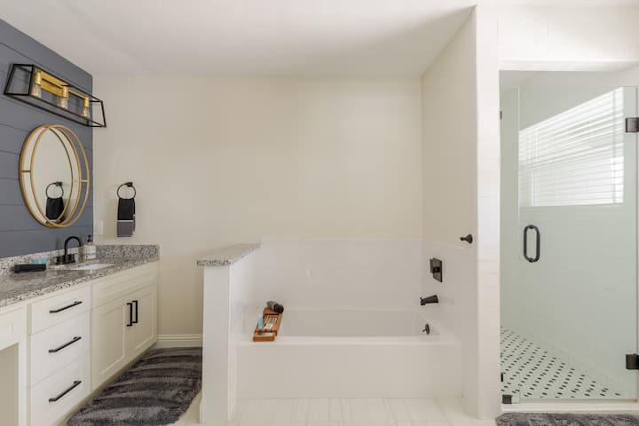 Une salle de bain moderne avec une baignoire blanche, un meuble-lavabo en granit, un miroir rond à cadre doré et une cabine de douche vitrée avec un sol en carrelage à motifs.