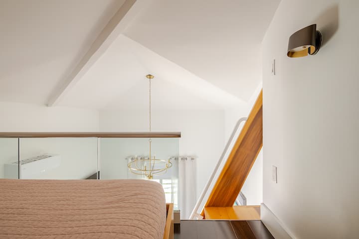 Un loft élégant avec un lit, une cloison en verre, un escalier en bois chaleureux et un lustre doré dans une pièce ensoleillée aux tons neutres doux.