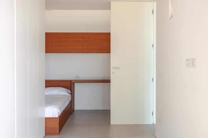 Une chambre minimaliste avec un lit simple, une tête de lit en bois et un bureau, partiellement visible à travers une porte coulissante blanche ouverte.