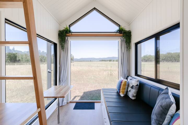 L'intérieur d'une tiny house lumineuse avec des plafonds voûtés, de grandes fenêtres et une vue sur une campagne ouverte avec des montagnes au loin.