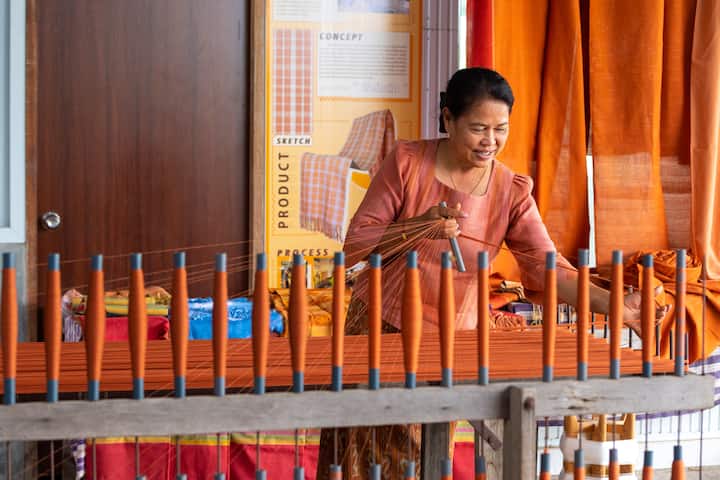 A person weaving fabric on a loom surrounded by orange and colorful textiles, showcasing traditional craftsmanship in Buriram, Thailand.