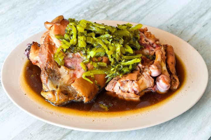 Close-up of a traditional Thai stewed pork leg served on a white plate, topped with pickled greens and surrounded by flavorful broth.