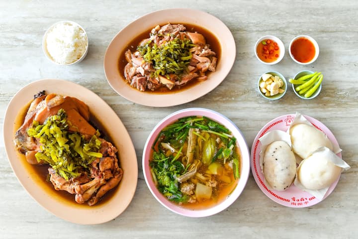 A spread of traditional Thai dishes including stewed pork leg topped with pickled greens, clear vegetable soup, steamed buns, rice, and various dipping sauces, served on a table.