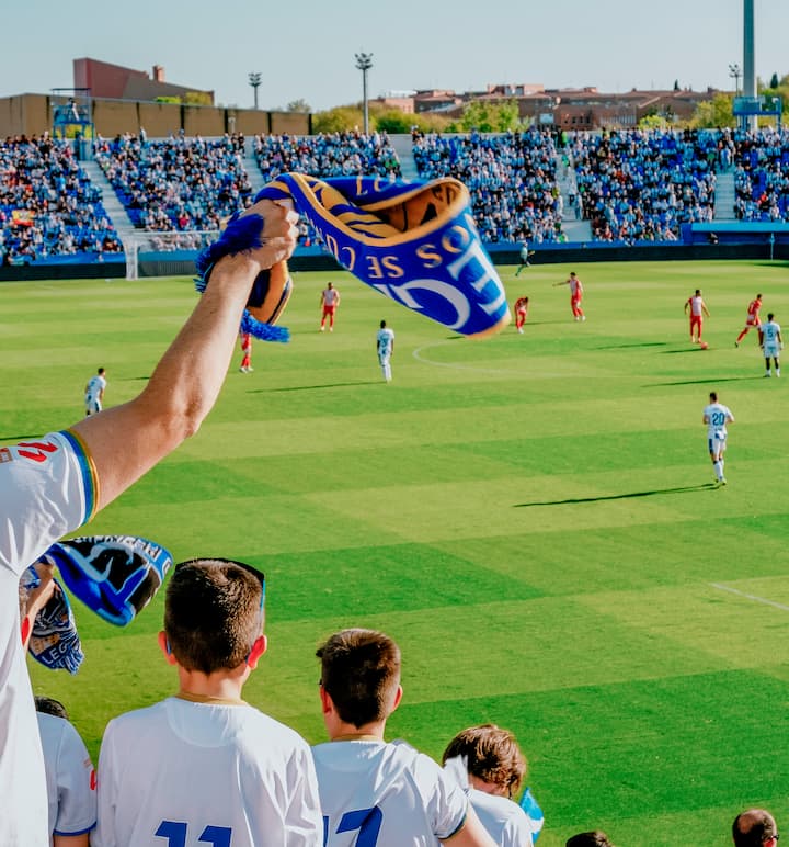 Un estadio repleto de aficionados, algunos con bufandas, que animan a su equipo durante un partido de fútbol bajo las luces del campo.