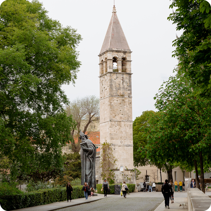 Une tour d'horloge en pierre se dresse au milieu d'arbres verdoyants dans un parc en plein air. Des personnes se promènent sur un chemin pavé à proximité et une grande statue se trouve au premier plan.