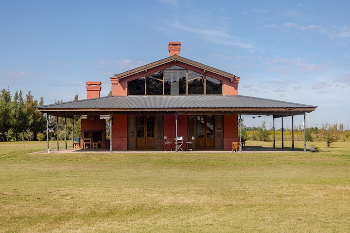 Une maison à la campagne avec un toit incliné, de grandes baies vitrées, des portes en bois et un porche ombragé, entourée de vastes prairies dans un cadre paisible.