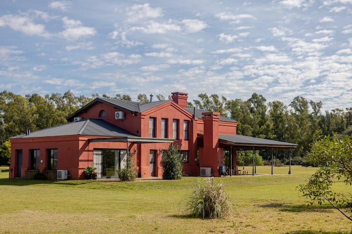 Une maison en briques rouges avec de grandes fenêtres, entourée d'une pelouse verte et d'arbres sous un ciel parsemé de nuages.