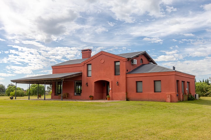 Une maison rouge de style champêtre avec un toit en métal, de grandes fenêtres et une terrasse couverte, entourée d'une pelouse verdoyante sous un ciel lumineux parsemé de nuages.