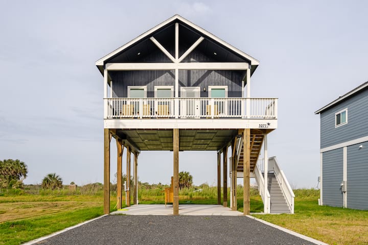 Maison moderne sur pilotis avec un toit triangulaire et de grandes fenêtres, située près d'une grande plaine et d'autres maisons identiques, idéale pour les séjours en bord de mer.