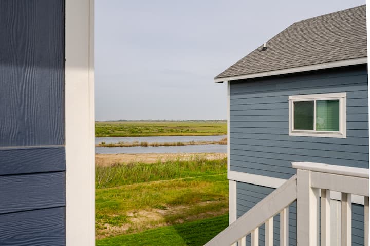 Vue d'une maison bleue avec une balustrade blanche surplombant un paysage verdoyant et une rivière paisible sous un ciel dégagé.