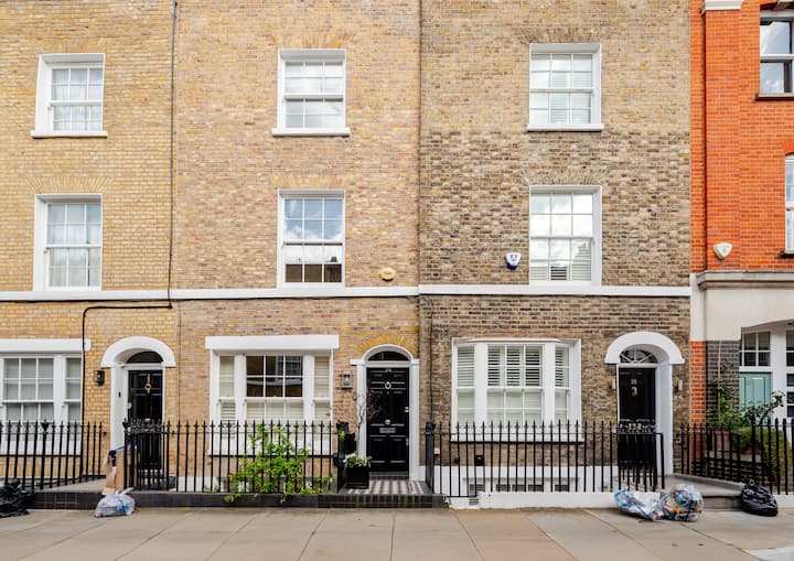 Rangée de trois maisons de ville en briques avec des clôtures en fer noir et de grandes fenêtres. Une plante pousse près de l'entrée centrale.