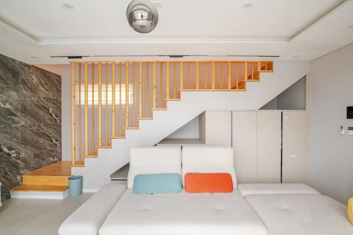 Un salon lumineux et ouvert avec un canapé blanc confortable, des coussins orange et bleus, un escalier en bois et un mur élégant en marbre gris.
