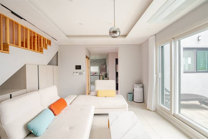 Un salon spacieux aux tons neutres, un canapé modulable, des coussins aux couleurs vives, un escalier et de grandes fenêtres laissant entrer la lumière naturelle.