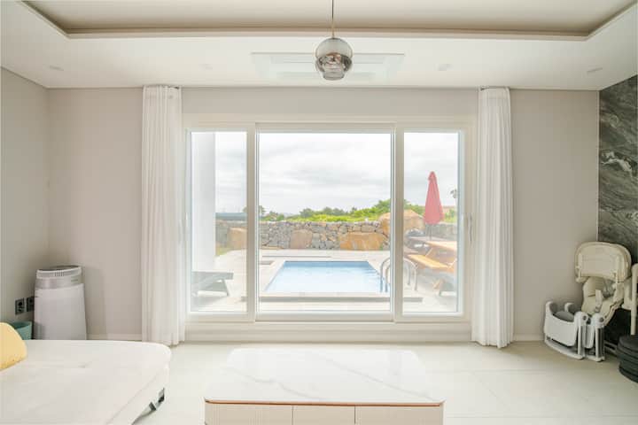 Un salon lumineux avec de grandes portes vitrées donnant sur une piscine, du mobilier d'extérieur et un mur en pierre entouré de verdure.