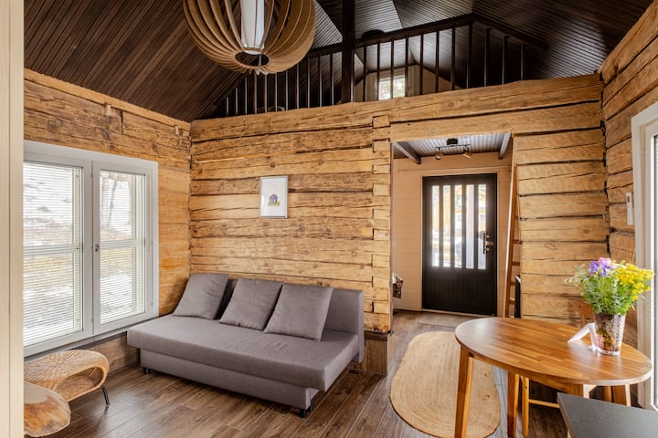 Une pièce chaleureuse dans un style cabane avec des murs en bois, un canapé gris, une table avec des fleurs coupées et une porte vitrée menant à l'extérieur.