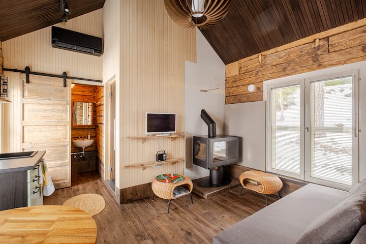 Intérieur d'une petite cabane mêlant éléments modernes et rustiques, notamment un plafond en bois, un foyer de cheminée noir, une télévision, des chaises en osier et une porte coulissante en bois menant à la salle de bain. Les stores des grandes fenêtres laissent entrevoir un paysage extérieur enneigé.