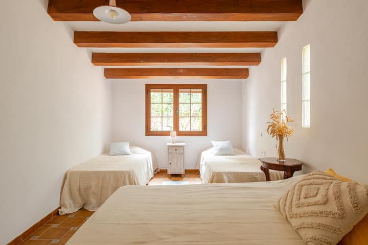 Chambre avec trois lits, des draps blancs, des poutres apparentes en bois et une fenêtre laissant entrer la lumière naturelle. La décoration de la pièce est minimaliste, avec un guéridon et des plantes séchées.