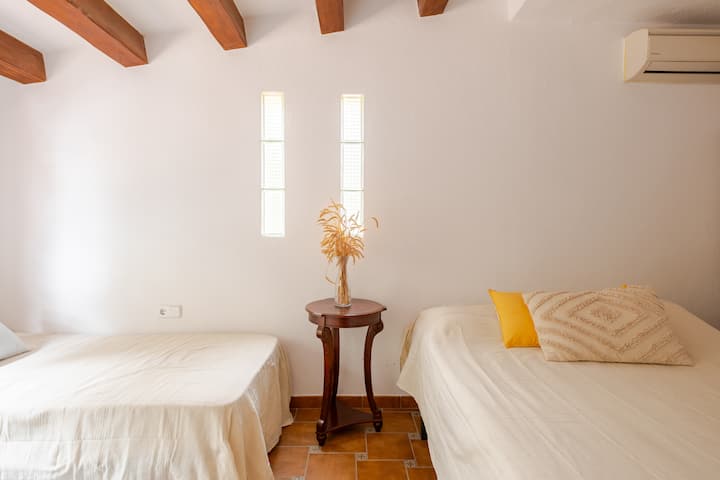 Une pièce lumineuse avec deux lits simples soigneusement faits avec des draps blancs, un guéridon avec des plantes séchées au centre et des poutres apparentes au plafond. La lumière pénètre à travers trois fenêtres étroites.