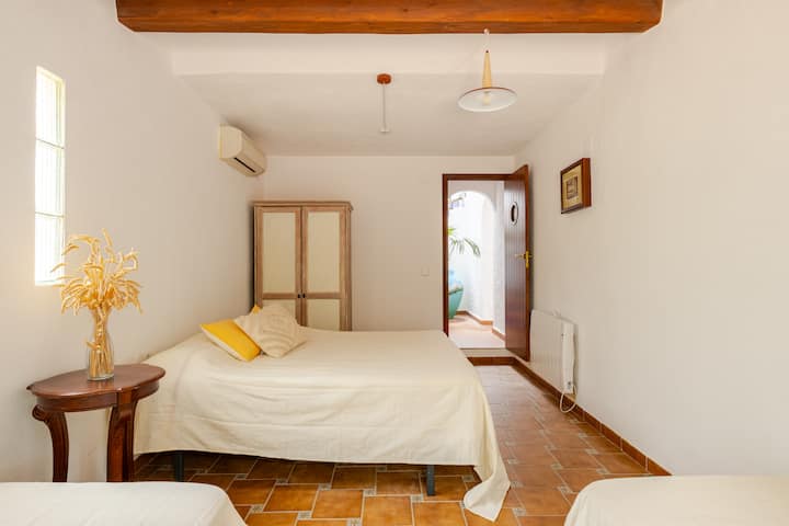 Une chambre accueillante avec du carrelage au sol, un lit double, des plantes séchées posées sur un guéridon en bois et une armoire. Une porte ouverte donne sur une terrasse avec des plantes luxuriantes.