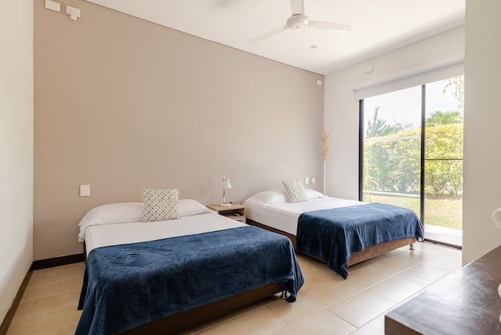 Une chambre bien éclairée avec deux lits jumeaux, des couvertures bleu marine, une décoration beige, un ventilateur de plafond et une porte vitrée coulissante donnant sur un jardin luxuriant.