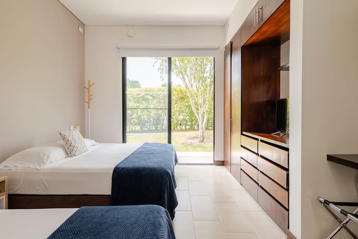 Une chambre minimaliste avec des meubles en bois, deux lits doubles et une porte vitrée coulissante offrant une vue sur un jardin ensoleillé et verdoyant.
