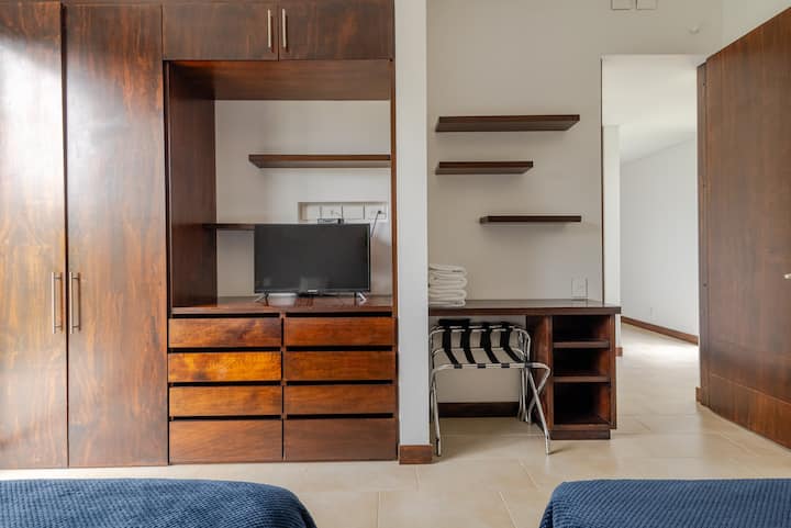 Une chambre avec une armoire en bois et des étagères, une télévision et un petit bureau avec une chaise. Une pile de serviettes pliées est posée sur le bureau.