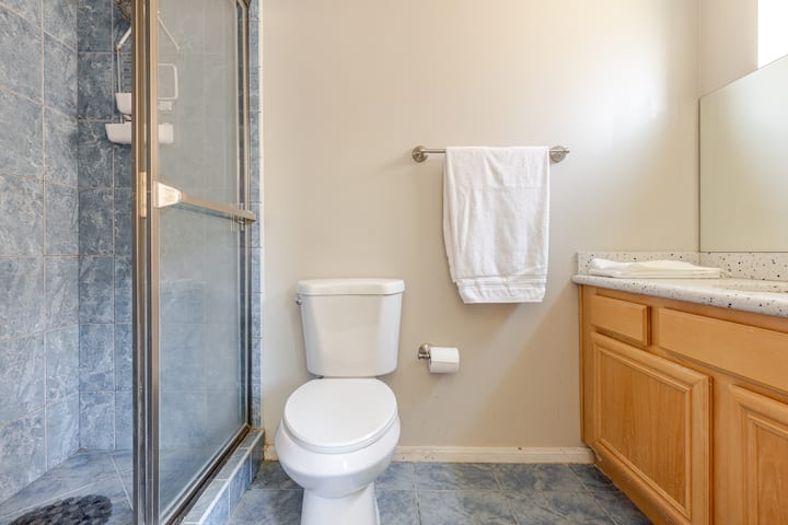 Salle de bain épurée avec une douche carrelée fermée par une porte en verre, des toilettes blanches, une serviette sur un porte-serviettes et une vasque mouchetée posée sur un meuble en bois.