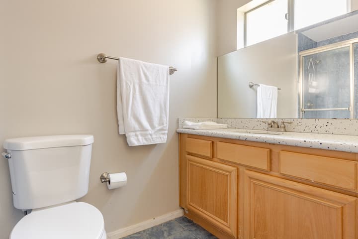 Salle de bain lumineuse avec des toilettes blanches, un meuble en bois clair sous une vasque mouchetée, un grand miroir et une serviette suspendue à un porte-serviettes argenté.