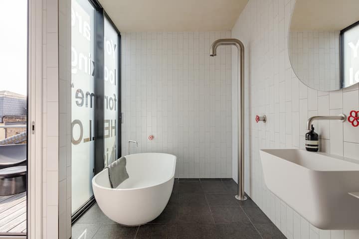 Salle de bain lumineuse avec une baignoire incurvée, un lavabo mural et des portes vitrées avec du texte visible. La pièce est dotée d'équipements modernes et les murs sont habillés de faïence blanche épurée.