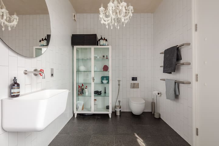Salle de bain lumineuse avec un miroir circulaire, des murs en faïence blanche, une vitrine en verre et des toilettes murales avec bidet.