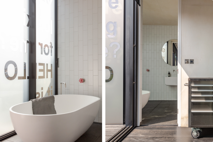 Vue côte à côte d'une salle de bain minimaliste avec une baignoire îlot à côté de fenêtres en verre dépoli et d'un deuxième espace avec un miroir rond au-dessus d'un lavabo, séparés par le cadre d'une porte coulissante.