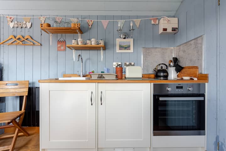 Une cuisine confortable avec des murs en bois bleu pastel, des placards blancs, un plan de travail en bois et des fanions décoratifs. On aperçoit également des tasses, une bouilloire, un grille-pain et un four.
