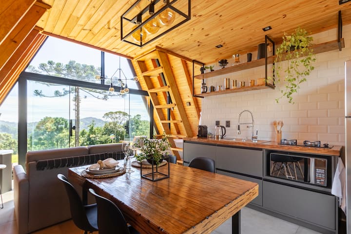 L'intérieur d'un chalet-tipi très lumineux avec une table à manger en bois rustique, une cuisine moderne avec des plantes retombantes et de grandes fenêtres offrant une vue panoramique sur les arbres et les montagnes.