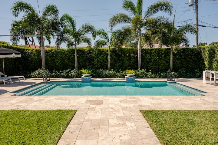 Une piscine d'un bleu translucide dans un jardin avec des palmiers verts, une haie bien entretenue, bordée de dalles en pierre et d'une pelouse.